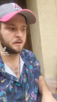 A close-up vertical video frame shows a light-skinned bearded man wearing a gray baseball cap with a pink brim and a large pink letter “P,” along with a blue floral-patterned button-up shirt. He faces slightly right, looking off-camera with a concerned or puzzled expression and his mouth slightly open. A doorway is behind him on the left, and a pale textured wall fills the right side. The lower-left corner contains the TikTok watermark and the text “TikTok” and “@saprious”. This meme is a video; captions are not available.