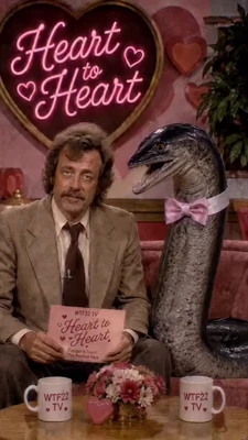 A vintage television talk-show set decorated in pink, with a large heart-shaped sign reading “Heart to Heart” in glowing cursive lettering, accompanied by three heart symbols. On the left, a mustached man with wavy brown hair sits on a couch in a tan suit, white shirt, and dark tie, looking toward the camera while holding a pink cue card. The card reads “WTF22 TV,” “Heart to Heart,” and smaller text including “Tania & Tonic” and “The …” beneath it. On the right, a tall gray dinosaur resembling a velociraptor sits upright beside him, wearing a pink bow tie and facing left with its mouth open. A leafy plant and heart-shaped wall decorations are behind them; a coffee table in front holds a flower arrangement, a pink heart ornament, and two white mugs labeled “WTF22 TV” with small heart symbols. This meme is a video; captions are not available.