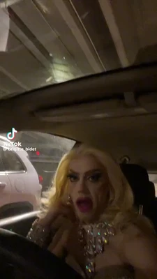 A vertical TikTok frame filmed inside a car in a dim parking garage: the ceiling beams and fluorescent lights are visible through the windshield, and a white SUV is parked alongside on the left. In the lower right, a blonde drag performer wearing heavy eye makeup, bright pink lipstick, a sparkling jeweled outfit, and a large rhinestone necklace looks toward the camera with wide eyes and an open mouth. The TikTok watermark and username “@brigitte_bid​​et” appear on the left. This meme is a video; captions are not available.