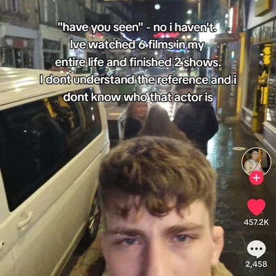 A vertical TikTok-style screenshot shows a young man with brown hair in the foreground, looking toward the camera with a tired or blank expression, seated beside the open window or side of a tram or bus traveling through a brightly lit city street at night. Other passengers are visible behind him, and wet pavement, storefronts, streetlights, and vehicles appear in the background. Large white text with black outlines across the upper half reads: `"have you seen" - no i haven't.` `live watched 6 films in my` `entire life and finished 2 shows.` `I dont understand the reference and i` `dont know who that actor is` On the right are TikTok interface elements: a circular profile picture above a pink plus sign, a heart icon with `457.2K`, and a comment bubble with `2,458`.