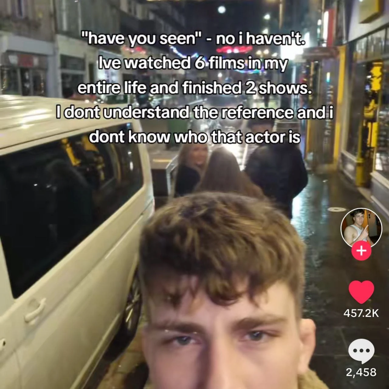 A vertical TikTok-style screenshot shows a young man with brown hair in the foreground, looking toward the camera with a tired or blank expression, seated beside the open window or side of a tram or bus traveling through a brightly lit city street at night. Other passengers are visible behind him, and wet pavement, storefronts, streetlights, and vehicles appear in the background. Large white text with black outlines across the upper half reads: `"have you seen" - no i haven't.` `live watched 6 films in my` `entire life and finished 2 shows.` `I dont understand the reference and i` `dont know who that actor is` On the right are TikTok interface elements: a circular profile picture above a pink plus sign, a heart icon with `457.2K`, and a comment bubble with `2,458`.