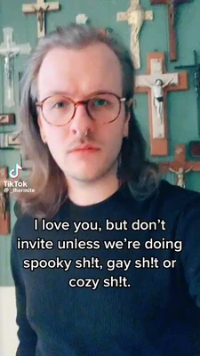 A person with long brown hair, round wire-frame glasses, and a black shirt faces the camera with a serious, unsmiling expression. They stand indoors against a teal wall covered with numerous crucifixes and crosses of different sizes, including several with corpus figures. Large white text with a black outline across the lower half reads: “I love you, but don’t invite unless we’re doing spooky sh!t, gay sh!t or cozy sh!t.” A TikTok watermark at lower left reads “TikTok” and “@_lhermite”. This meme is a video; captions are not available.