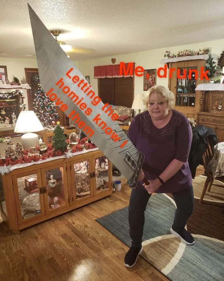 A woman with short blond hair stands in a decorated living room, holding a large gray triangular object diagonally across her body from the upper left toward the lower right; dark mechanical details are visible near its lower end. She wears a dark purple top, dark pants, and black slip-on shoes, with her legs spread apart and a neutral expression. The room contains a Christmas tree, holiday decorations, a lit table lamp, a wooden display cabinet, furniture, and a patterned rug. Large red text across the top reads “Me drunk,” and diagonal red text across the gray object reads “<Letting the homies know I love them”.