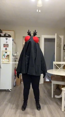A person stands centered in a kitchen, facing away from the camera and covered by a loose black, cape-like costume that reaches from the shoulders to the knees. Two large red circular shapes sit near the top of the costume, with black antler-like decorations rising above them; black-gloved hands hang at the sides, and the person wears black pants and lace-up boots. A white refrigerator with magnets and papers stands on the left, an open doorway is behind the person, and a white table and crossed-back chair are on the right. A ceiling light hangs overhead. In the lower-left corner, the watermark text reads exactly: “TikTok” and “@walmartmothman”. This meme is a video; captions are not available.