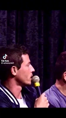 A vertically framed video with black bars at the top and bottom shows two men onstage against dark purple curtains. In the foreground, a dark-haired man in left profile wears a black jacket with white trim over a white shirt and holds a handheld microphone with a yellow band near his mouth, looking toward the man beside him. The second man is partially visible on the right, wearing a purple shirt and facing right. A TikTok watermark on the left reads “TikTok” and “@sofunny369”. This meme is a video; captions are not available.