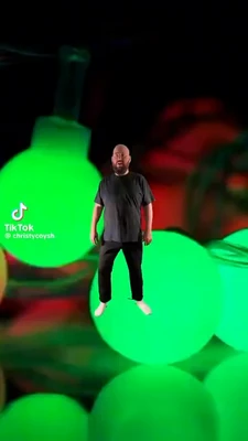 A bald, bearded man in a dark gray T-shirt and black pants stands centered, appearing to float above or in front of a brightly glowing neon-green sphere, with his arms hanging at his sides and a neutral expression. The dark background is filled with large blurred green, red, and orange rounded shapes resembling glowing game or toy objects. In the lower-left corner, white TikTok branding reads “TikTok” and “@christycoysh.” This meme is a video; captions are not available.