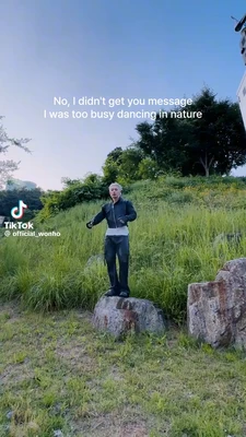 A vertical TikTok frame shows a light-haired young man standing on a large rock in a grassy, overgrown outdoor area, with his arms bent and raised slightly as if dancing. Trees and shrubs fill the background beneath a clear blue sky, and part of a building appears along the upper-right edge. White text near the top reads, “No, I didn't get you message / I was too busy dancing in nature”. The TikTok logo and the text “TikTok” and “@official_wonho” appear in the lower-left corner. This meme is a video; captions are not available.