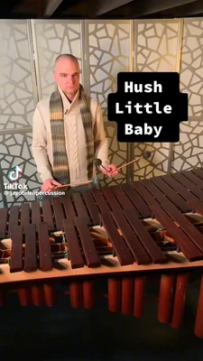 A bald man in a cream cable-knit sweater and long striped scarf stands behind a large wooden marimba, holding two mallets over its rows of dark wooden bars; his face is serious and he looks toward the camera. The marimba fills the foreground, with reddish resonator tubes visible beneath the bars. Behind him is a folding screen with an irregular geometric pattern, and dark ceiling beams are visible above. A black stacked caption near the upper right reads exactly, “Hush Little Baby.” In the lower-left corner, the TikTok logo and watermark read exactly, “TikTok” and “@jayobrienpercussion.” This meme is a video; captions are not available.