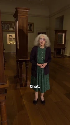 In a dimly lit museum gallery, an older woman with shoulder-length gray hair and glasses stands centered in front of a tall wooden display cabinet, facing the camera with her mouth open as if speaking and her hands clasped at her waist. She wears a dark cardigan over a green patterned dress and black shoes. Other wooden display cases, framed paintings, and exhibits line the room. White caption text at the bottom reads, “Chat,”. This meme is a video; captions are not available.