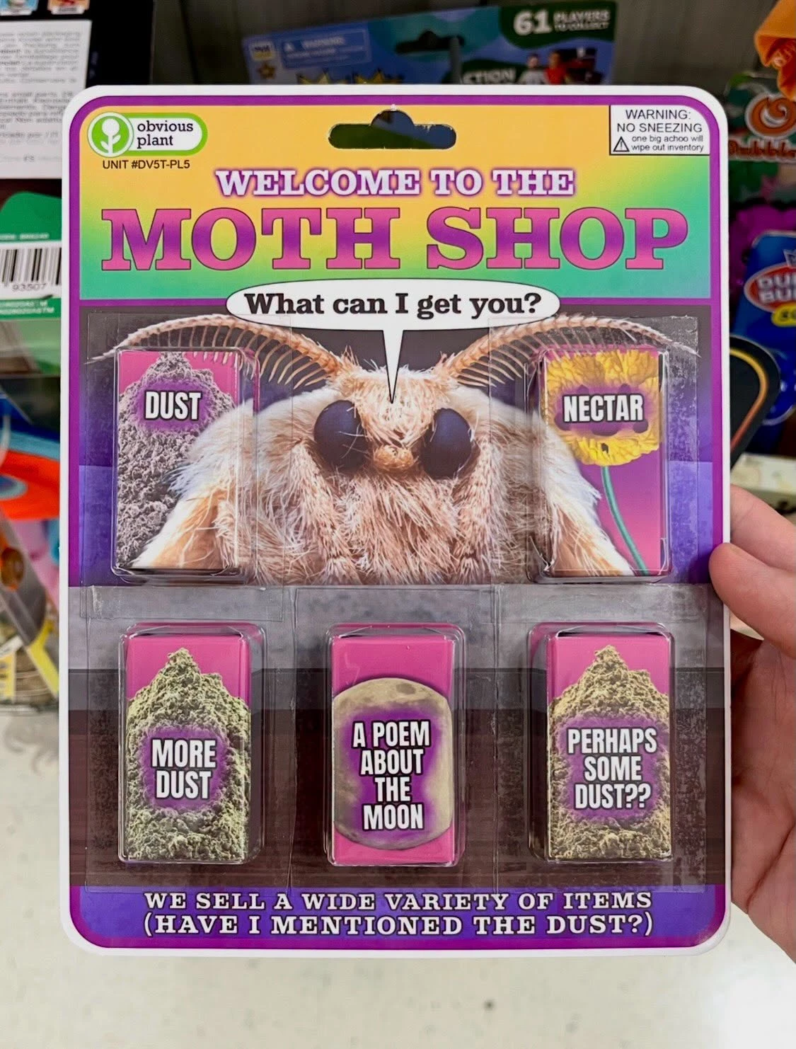 A colorful packaged novelty product is photographed in a store, with a large fuzzy moth pictured in the center, its wings spread behind its head, antennae extending upward, and large dark eyes facing forward. The package has a green-and-white “obvious plant” logo at upper left, the text “UNIT #DV5T-PL5,” and a warning label at upper right reading “WARNING: NO SNEEZING” and “one big achoo will wipe out inventory.” Large pink-and-white lettering says “WELCOME TO THE MOTH SHOP,” with a speech bubble below reading “What can I get you?” Two upper compartments are labeled “DUST” and “NECTAR,” and three lower compartments contain items labeled “MORE DUST,” “A POEM ABOUT THE MOON,” and “PERHAPS SOME DUST??” The bottom banner reads “WE SELL A WIDE VARIETY OF ITEMS (HAVE I MENTIONED THE DUST?)” Shelves of other merchandise are visible in the background, and a hand holds the package along the right edge.