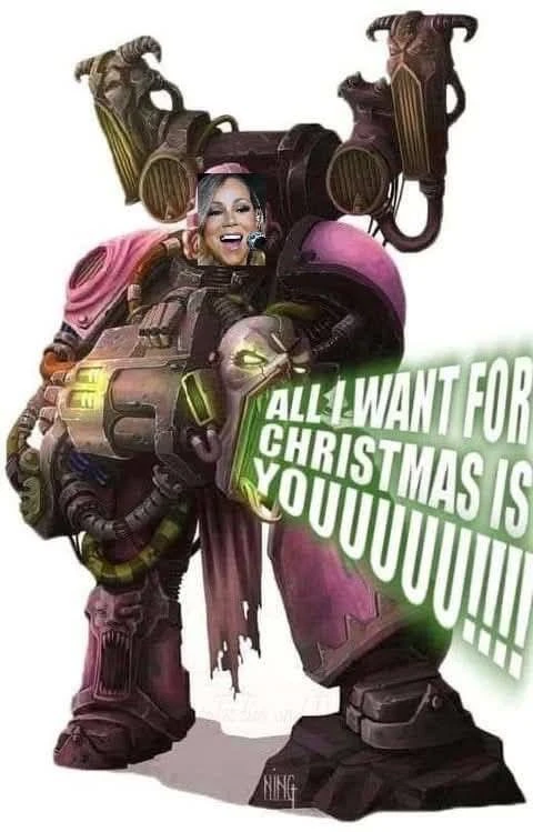 A vertically oriented meme on a white background depicts the heavily armored Borderlands character Krieg, with Mariah Carey’s smiling, open-mouthed face superimposed over his head. He stands facing forward in bulky brown, black, and purple armor, carrying a large weapon across his body; curved, horn-like equipment rises behind his shoulders, and his gloved hands grip the weapon near the center. A tilted green sign covers the lower-right portion of the figure and contains large white uppercase text reading: “ALL I WANT FOR CHRISTMAS IS YOUUUUUUUUUUUUUUUUUUU.”